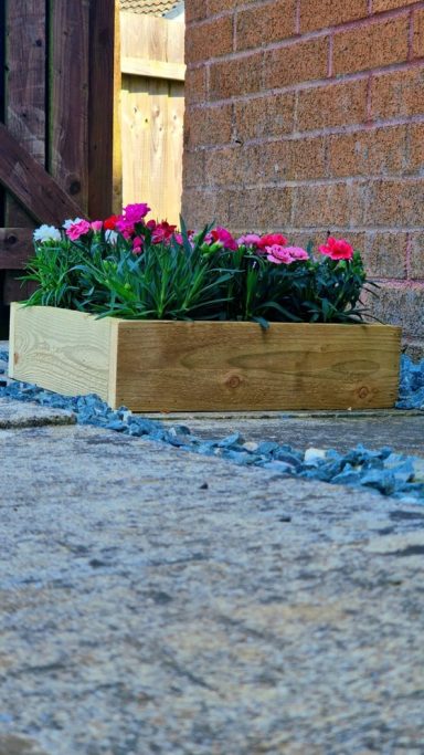 Wooden planter box filled with pink and white flowers, set alongside stone pathway.